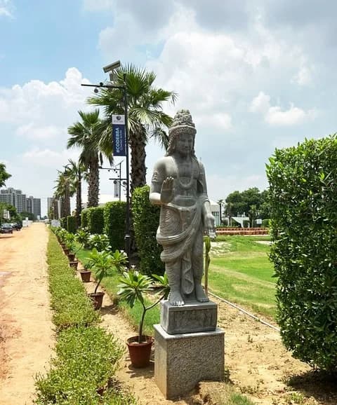 Standing stone sculpture on a plinth beside the planted avenue at the site, flanked by clipped hedges, palms and a lamp post carrying project banners.