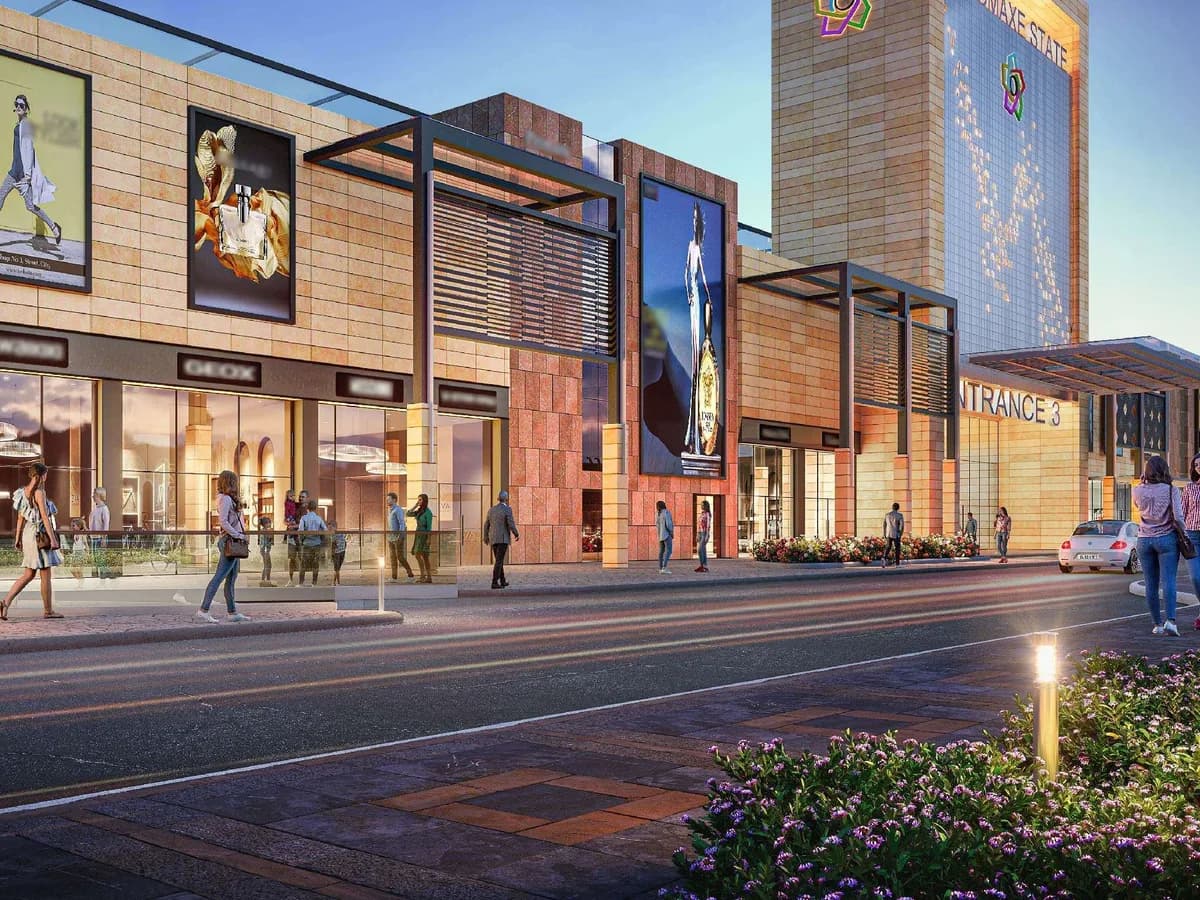 Evening render of the Shopping District street frontage with lit shopfronts, billboards and a tower branded The Omaxe State beside Entrance 3